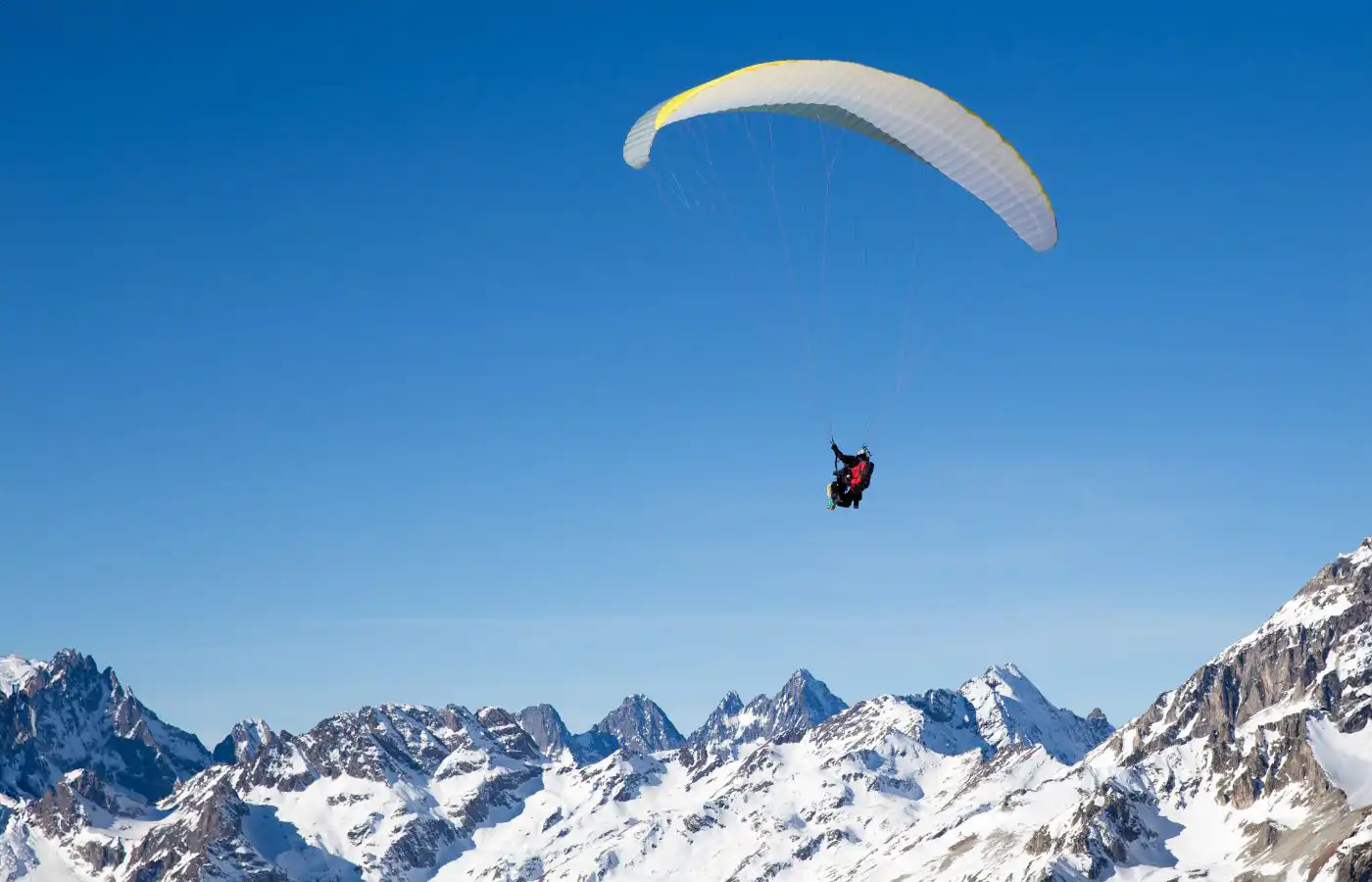 Personne en parapente survolant des montagnes enneigées sous un ciel bleu clair, avec des sommets alpins en arrière-plan.