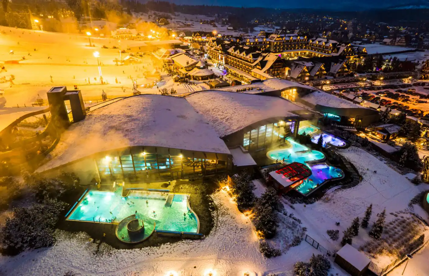 Vue aérienne d'un complexe thermal en montagne, entouré de neige avec des piscines chauffées et des bâtiments traditionnels en arrière-plan.