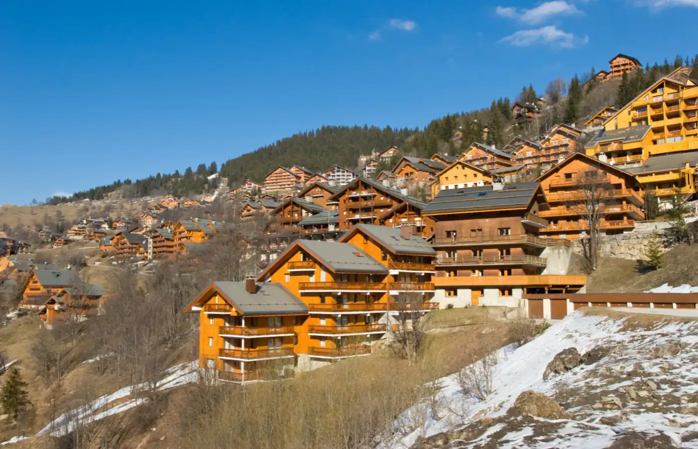 Vue d'un ensemble de chalets et d'appartements en bois dans une station de ski de montagne sous un ciel bleu, avec des pentes partiellement enneigées.