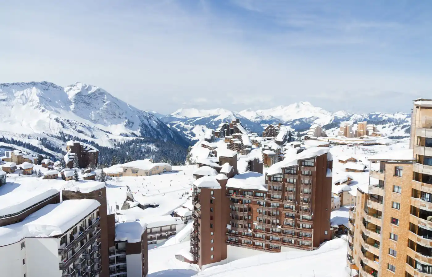 Vue panoramique de bâtiments d'appartements dans une station de ski entourée de montagnes enneigées sous un ciel bleu clair.